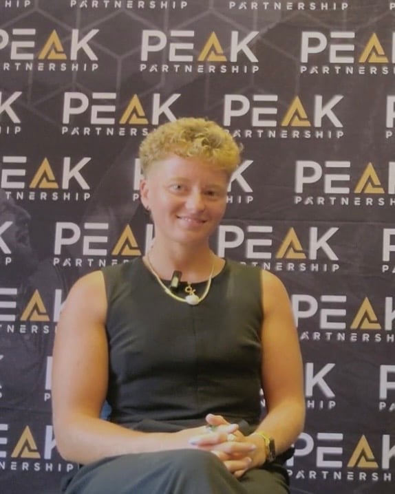 Person with short yellow hair wearing a black sleeveless shirt and gold chain necklace smiling at a PEAK Partnership branded event backdrop